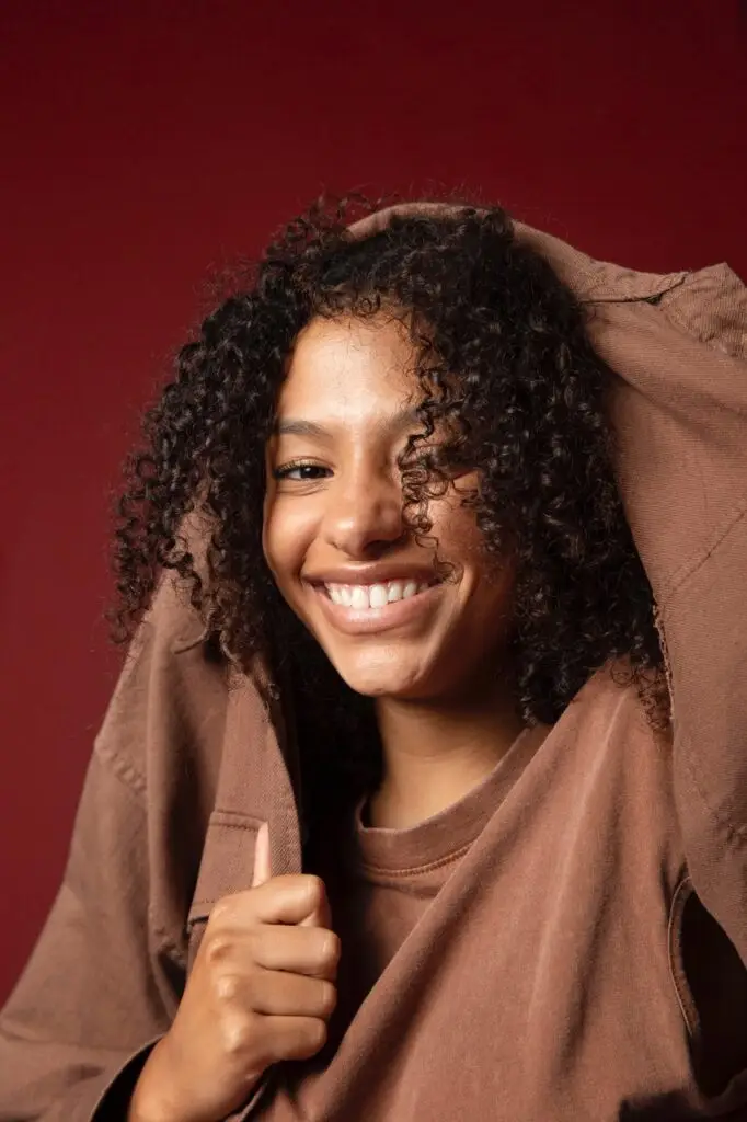 Jeune femme souriante avec des cheveux bouclés, portant un sweat à capuche marron, posant devant un fond rouge.