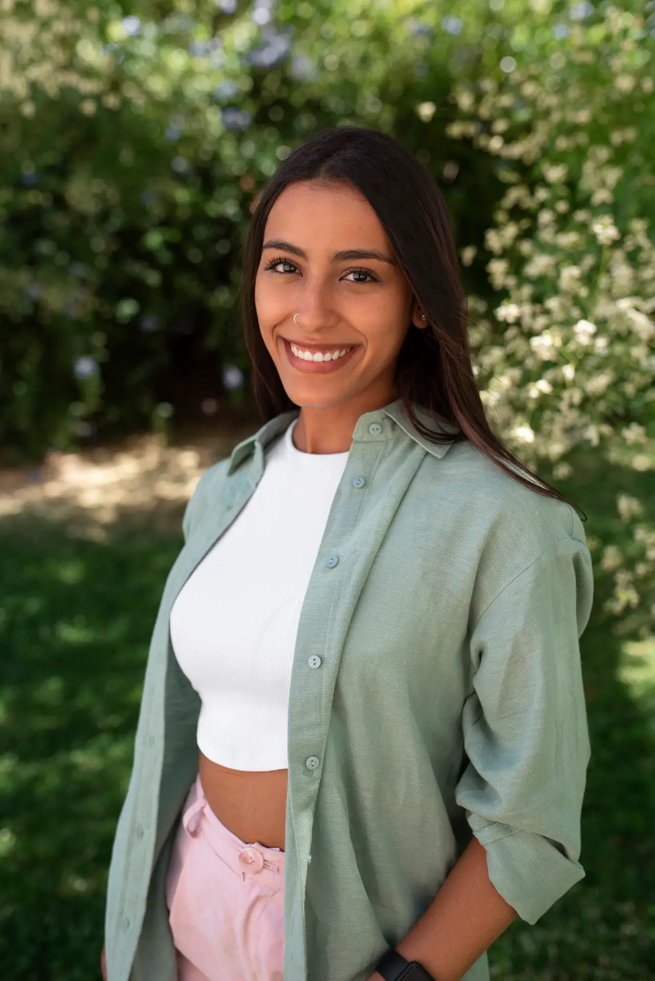 Jeune femme souriante avec une chemise verte et un top blanc posant dans un jardin verdoyant, avec des arbres et des fleurs en arrière-plan.