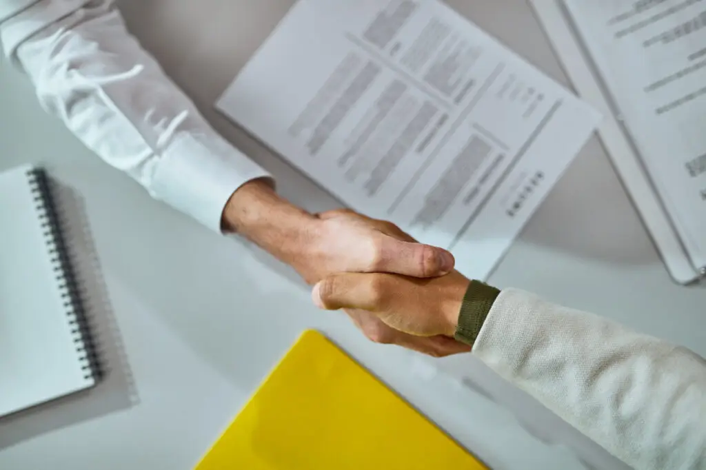 Deux personnes se serrant la main au-dessus d'une table avec des documents et des CV, symbolisant la fin d'un entretien d'embauche.