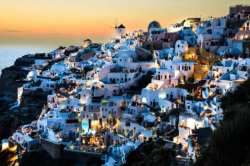 Vue nocturne des maisons blanches illuminées sur les falaises de Santorin en Grèce, avec des moulins à vent et un coucher de soleil en arrière-plan.