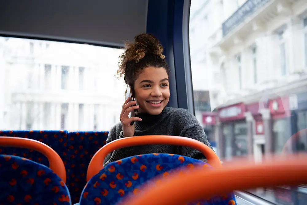 Jeune femme souriante avec des cheveux bouclés parlant au téléphone portable, assise dans un bus avec des sièges colorés, regardant par la fenêtre sur une rue animée.