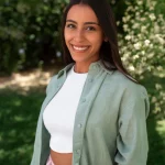 Jeune femme souriante avec une chemise verte et un top blanc posant dans un jardin verdoyant, avec des arbres et des fleurs en arrière-plan.